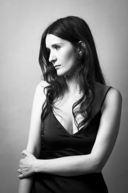 A white woman with long brown hair and a slender frame, she looks off to one side, one arm around her waist gripping the other arm.