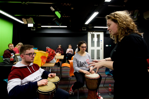 A workshop leader is supporting a young Disabled musician with a percussion instrument similar to bongos