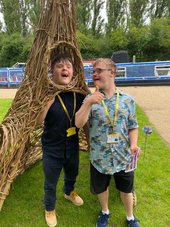 Two young disabled men smile and pose in a small willow hut, one with his head popping out through a hole.