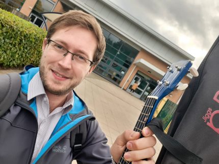 A white man in front of a school holds up a guitar and smiles as he takes a selfie