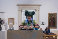 The artist Rebekah Ubuntu is DJ-ing on a sparkling turquoise audio mixer. They have braided cyan coloured hair and wear a gold sequinned jacket and colourful light-up rings on their fingers. Behind them are paintings on a white gallery wall.