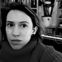 A black and white photograph of a white woman with brown hair. She’s wearing a ribbed, high neck jumper. In the background is a studio space with a keyboard and shelves on the wall.