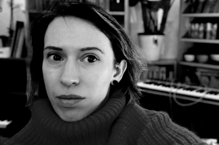 A black and white photograph of a white woman with brown hair. She’s wearing a ribbed, high neck jumper. In the background is a studio space with a keyboard and shelves on the wall.