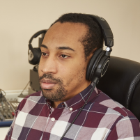 A portrait photograph of Andre Louis, a brown-skinned man with short black hair wearing black headphones and a checked shirt.