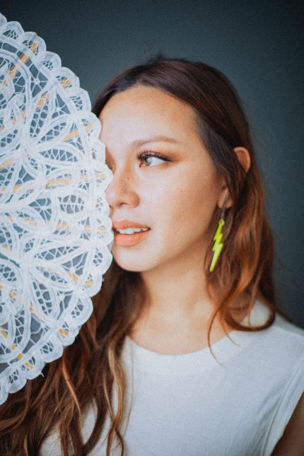 Photograph of Sha, an East Asian woman in her 30s, holding a white fan. She has long dark brown hair and is wearing a white top with a neon yellow lightning bolt earing.