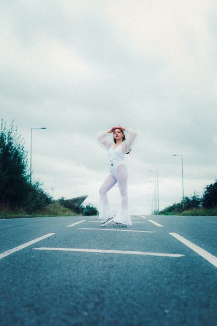 Photograph of Sha, an East Asian woman in her 30s, standing on a road against a cloudy sky. She has long dark brown hair and is wearing all white, flared polkadot see-through bottoms, a white organza fluffy top and roller skates.