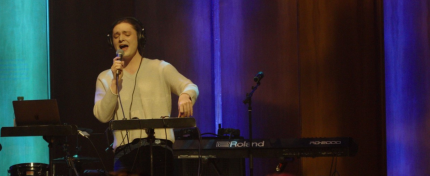Geo Aghinea performing on stage. They are wearing headphones and are singing into a microphone, whilst using a laptop and other audio equipment,.