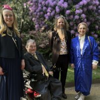Picture of the Drake Music team in front of a back drop of shrubs and purple bushes. Sally Currie, Tim Yates and Deborah Borg Brincat are standing, with John Kelly sitting in his wheel chair. The team are in smart attire, and are smiling at the camera.