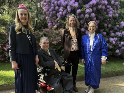 Picture of the Drake Music team in front of a back drop of shrubs and purple bushes. Sally Currie, Tim Yates and Deborah Borg Brincat are standing, with John Kelly sitting in his wheel chair. The team are in smart attire, and are smiling at the camera.