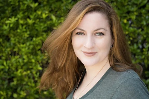 A headshot of Ingrid Plum against a background of green leaves. Ingrid is wearing a grey t-shirt and is smiling at the camera.