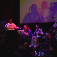 Photograph of Muminah, Reese Quinlan, Miss Jacqui and L U C I N E performing on stage. Reese and Miss Jacqui are sitting in the middle, performing vocals. Muminah and L U C I N E are stood either side, with L U C I N E playing bass. The stage is set in a purple colour, with graphics of the artists behind on a screen.