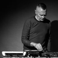 A black and white photograph of Graham using audio equipment and an iPad against a dark back drop. Graham is looking down at the equipment on the desk, and has an expression of concentration on his face.