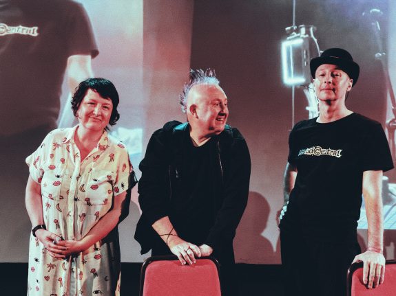A photograph of (left to right) Nicola Woodham, Lee Holder and zen olenski. The three are standing on stage, leaning on red chairs in front. Nicola and zen are looking at the camera, smiling, whilst Lee is looking to their left.