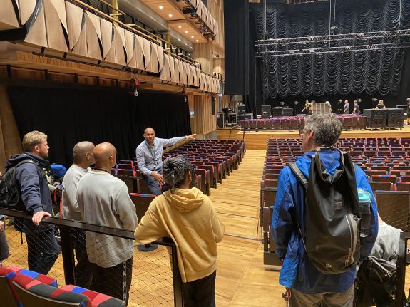 A group of people on a tour of a large, bright concert hall. They are standing towards the back, and are looking at the stage which is being set.