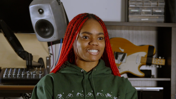 A screengrab of Sarah Adedeji during an interview. Sarah has red braided hair, and is looking towards the direction of the interviewer. She is smiling.
