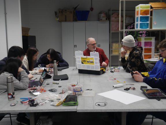A group of young students and their teacher sat around a desk. They are working with a laptops and electrical components.