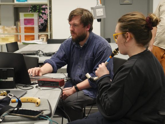 A participant with a ceramic wind instrument raised to their lips, whilst another participant is working on a laptop.