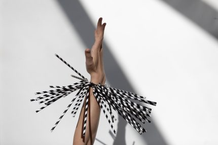 A hand and arm pointing straight upwards against a light, white background. Around the wrist is a bracelet of white and black straws pointing in different directions.