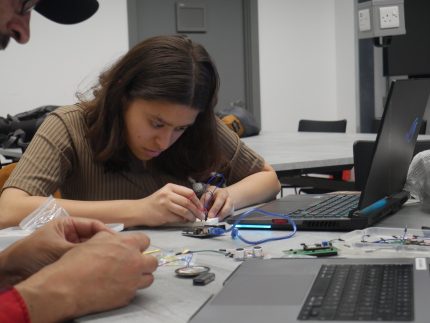 A hackathon participant working with electrical components and a laptop on a desk. They have a concentrated look on their face.