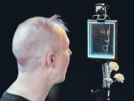 The back of zen's head facing the _PhotoSYNTH 4D_ with their mouth open. The instrument is mounted, with cameras and sensors on top of a tablet screen, and flowers attached below.