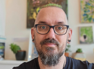 A white, green haired man smiling. He is wearing glasses, hearing aids, has a beard and is sitting in a gallery.