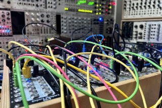 A close-up of a modular synthesizer with multiple brightly colored patch cables (yellow, pink, green, black) connecting various modules. The modules feature rows of knobs, buttons, and LED lights glowing blue, red, and pink. A wooden case holds the modules at an angle, with a larger modular synthesizer visible but blurred in the background.