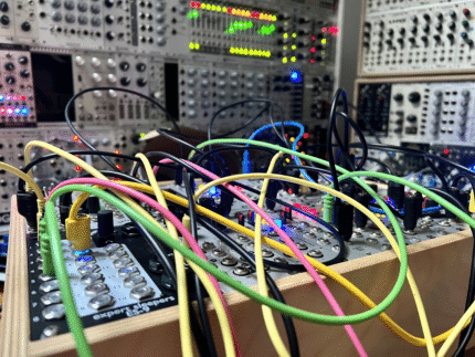 A close-up of a modular synthesizer with multiple brightly colored patch cables (yellow, pink, green, black) connecting various modules. The modules feature rows of knobs, buttons, and LED lights glowing blue, red, and pink. A wooden case holds the modules at an angle, with a larger modular synthesizer visible but blurred in the background.