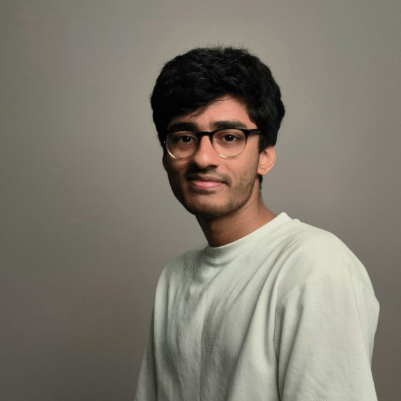 A man in his late twenties with short dark hair, glasses, and a white t-shirt, smiling slightly against a soft grey background.