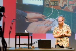A man with glasses, orange hair and a long beard standing at a desk, presenting. He has a laptop and a small instrument on a stool, with a lamp overhead.
