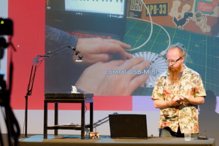 A man with glasses, orange hair and a long beard standing at a desk, presenting. He has a laptop and a small instrument on a stool, with a lamp overhead.