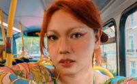 A woman with red hair sits on a bus, framed by yellow-and-blue seats. She wears bold pink-and-green makeup and a psychedelic patterned top in bright colours. Lightning-bolt and pigeon earrings dangle as she looks straight into the camera with a calm, steady gaze.