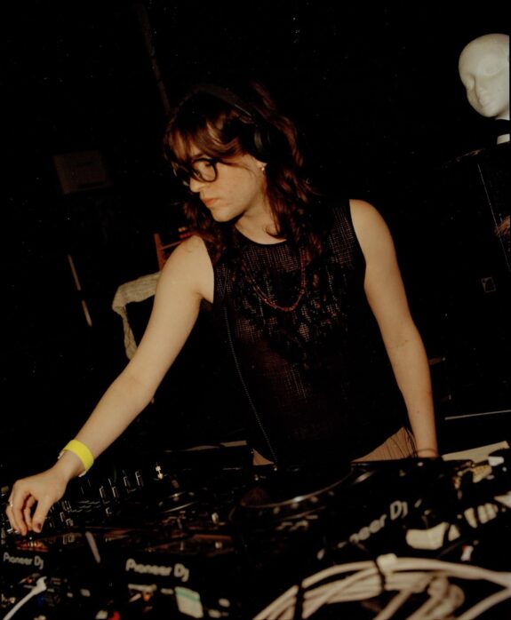 A young person with dark hair and glasses mixing on DJ decks in a nightclub. They are wearing a black vest.