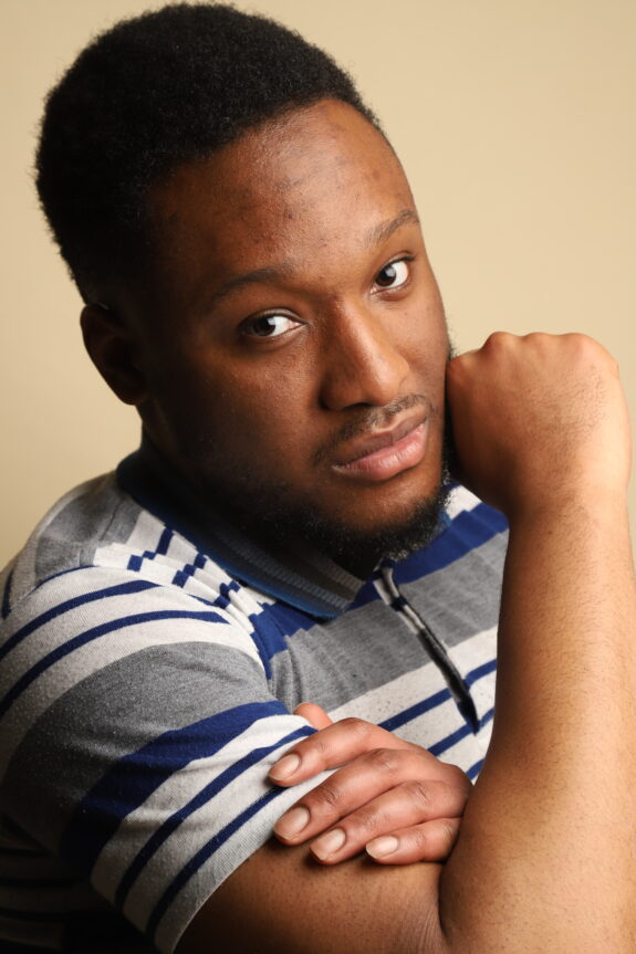 A close-up of a young Black man looking at the camera. He has a serious expression on his face, and is resting his face against his right hand.