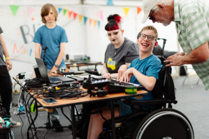 A young boy sat at a desk in a wheelchair in a large, white festival tent. He is looking at an older man to his left, smiling. On the desk is lots of audio equipment: MIDI keyboards, sample pads, a Mac and cables. A facilitator is sat to his right.