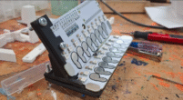 A grey and black rectangular-shaped device with 2 rows of silver thumbprint shaped sensors. It sits on a messy workbench.
