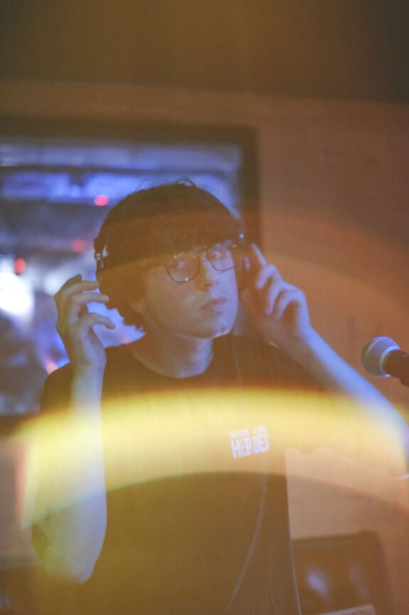 A young man wearing a hat, looking at the camera. He is about to take off headphones in front of a microphone, with a big lens flare in bottom of the image.