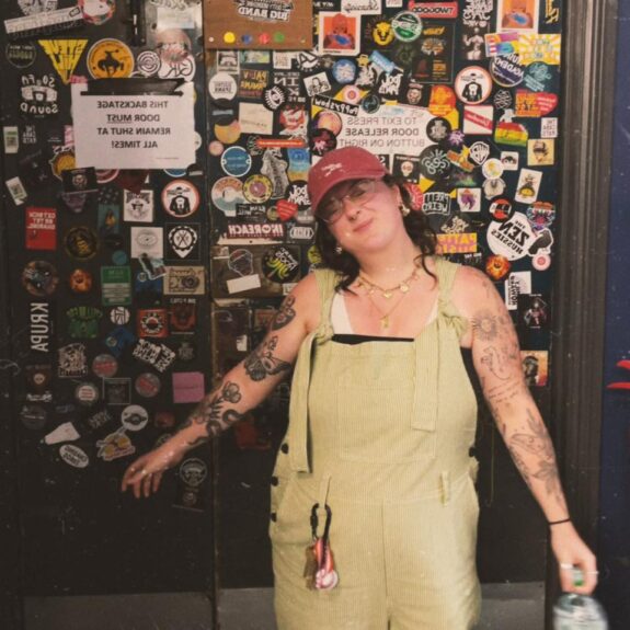 A white woman with shoulder length brunette hair underneath a red cap and tattoos on her outstretched arms.