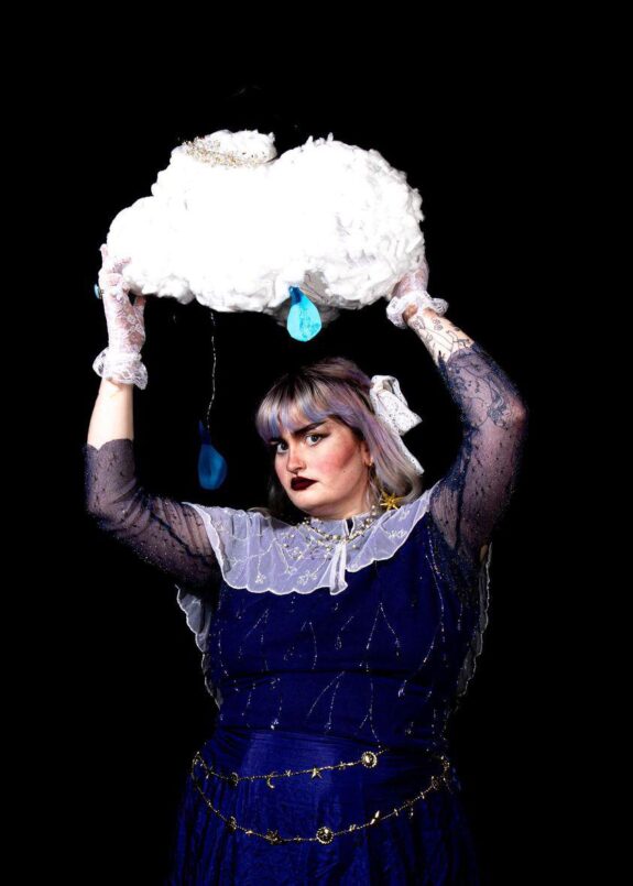 A young person in Gothic-style clothing holding a fluffy cloud above their head. They are looking at the camera with a serious expression. Their hair is a silver fringe, and they wear dark lipstick.
