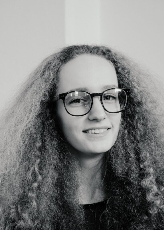 A black and white portrait photograph of a person with long, voluminous, curly hair, wearing glasses and smiling at the camera with a relaxed and friendly expression.