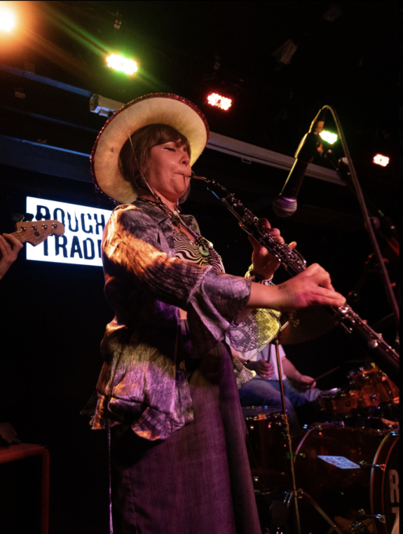 Pea, a young musician wearing a cowboy hat plays cor anglais into a microphone on stage alongside a drummer and bassist. An illuminated ‘Rough Trade’ sign is visible in the background.