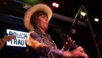 Pea, a young musician wearing a cowboy hat plays cor anglais into a microphone on stage alongside a drummer and bassist. An illuminated ‘Rough Trade’ sign is visible in the background.