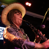 Pea, a young musician wearing a cowboy hat plays cor anglais into a microphone on stage alongside a drummer and bassist. An illuminated ‘Rough Trade’ sign is visible in the background.