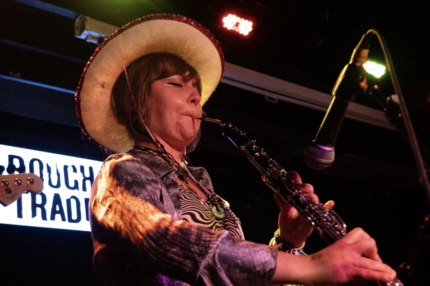 Pea, a young musician wearing a cowboy hat plays cor anglais into a microphone on stage alongside a drummer and bassist. An illuminated ‘Rough Trade’ sign is visible in the background.