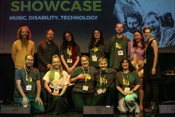 A group photograph of the DM team on-stage. They form two lines of 5 people; the back standing and the front kneeling. They are smiling, and are mostly wearing grey and yellow t-shirts with the Drake Music logo in the centre.