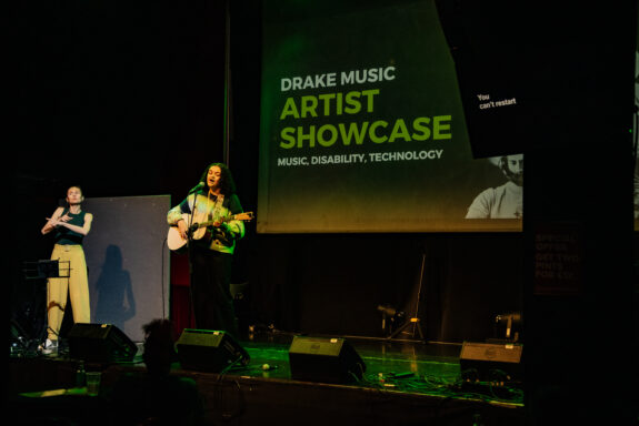 A musician, Jasmine Marie, standing on stage with an acoustic guitar in her hands. She is smiling and singing into the microphone. Behind is a large screen with yellow and white text that reads 'Drake Music Artist Showcase. Music, Disability, Technology'.