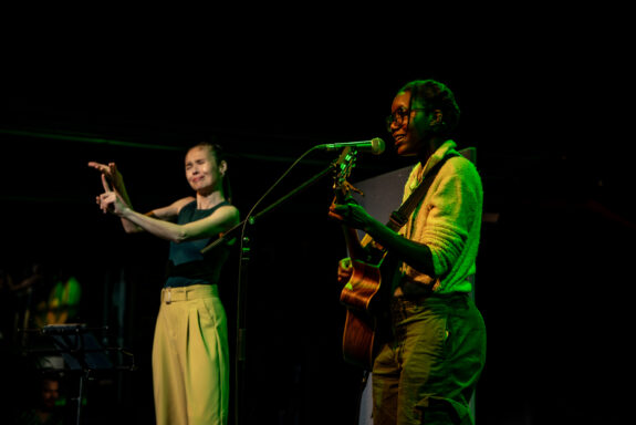 A musician, Dani Osoba, on stage with her guitar. Dani is smiling softly, and is singing into a microphone. The light is set to green.
