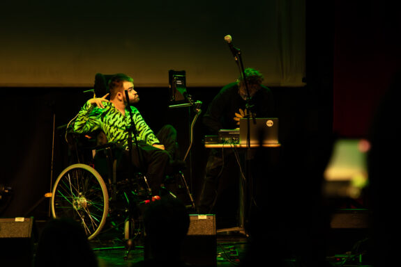 A musician, The Shouting Mute, on-stage in his wheelchair and with his communication aid in front of him. He is smiling, and wears a zebra patterned jacket. The lighting is a soft green.