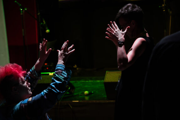 A side-profile shot of two people signing with their hands to each other. One person has redish pink hair and glasses. The other person is wearing a black top and has dark hair.