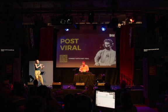 A DJ, Post Viral, sitting on-stage at his decks. His hands are outstretched as he is mixing. There is a microphone in front of him which he is speaking into.