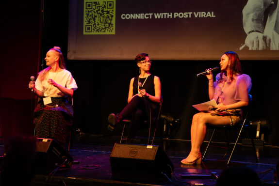 Sha Supangan, Sally Currie and Lisa Heywood on-stage. They are sitting down and speaking into microphones. They stage lighting is a soft pink.